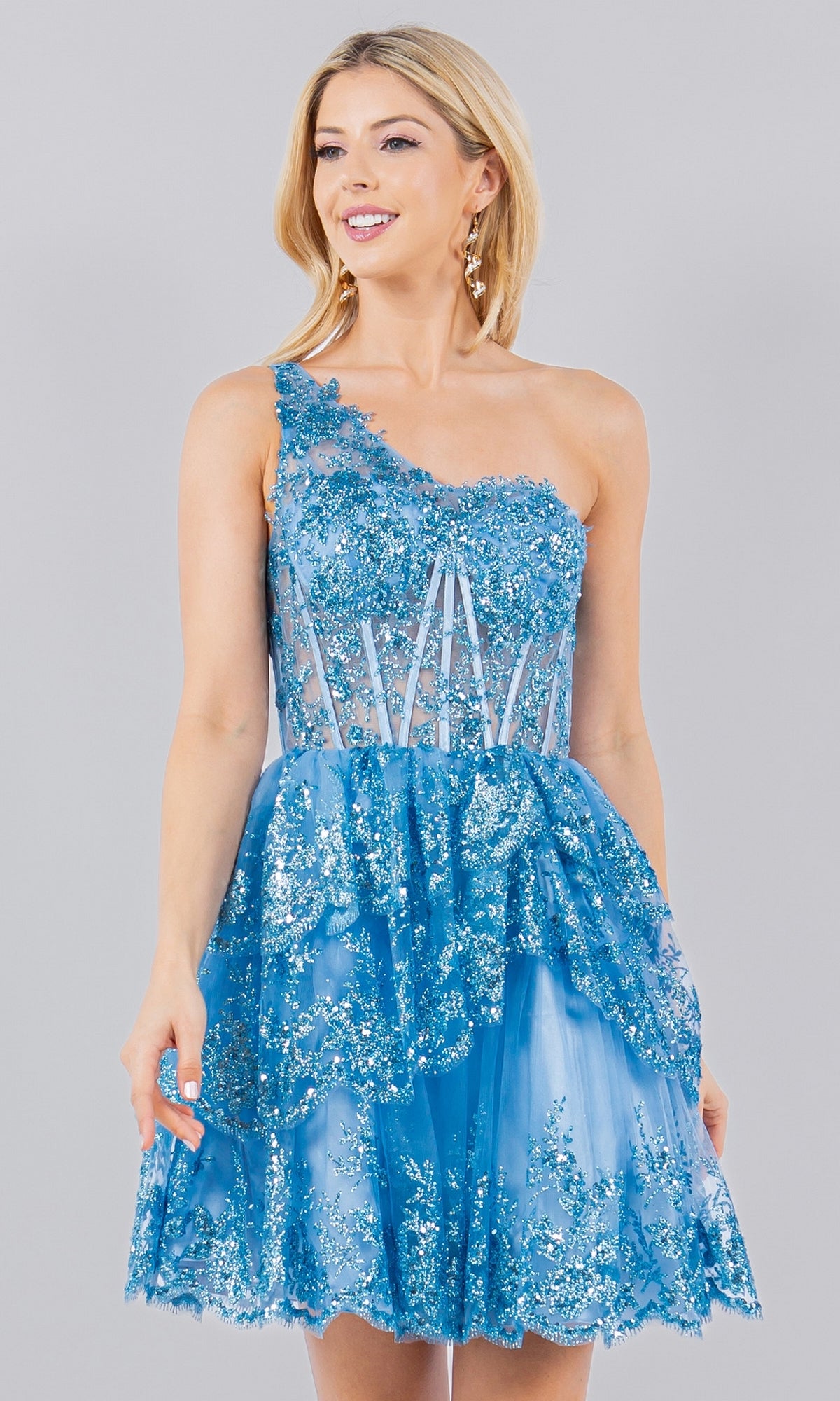 A woman models the One-Shoulder Short Tiered Homecoming Dress 5132J, featuring blue floral lace details and sheer sections, standing against a plain gray background.